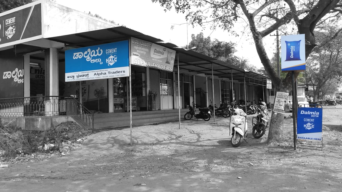 Cement dealer shopfront with a Dalmia branded fascia board, pole-mounted sign and forecourt standee in daylight.