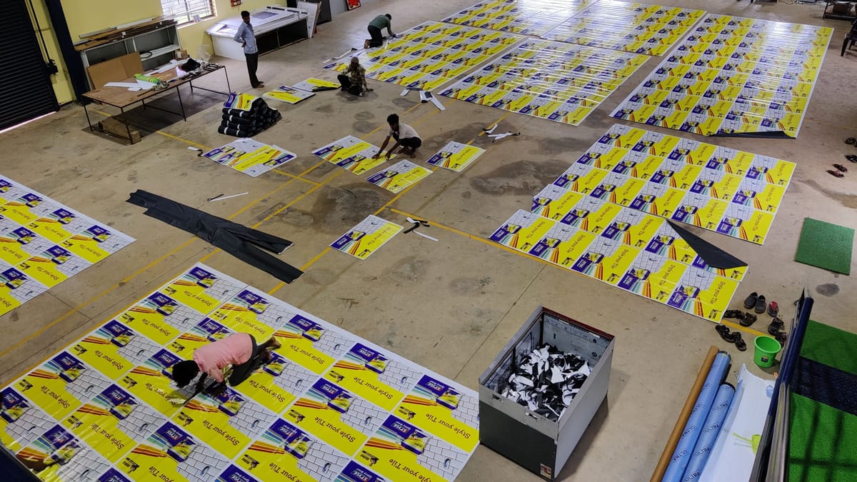 Overhead view of a printing workshop floor covered with yellow printed vinyl panels as four workers trim them.