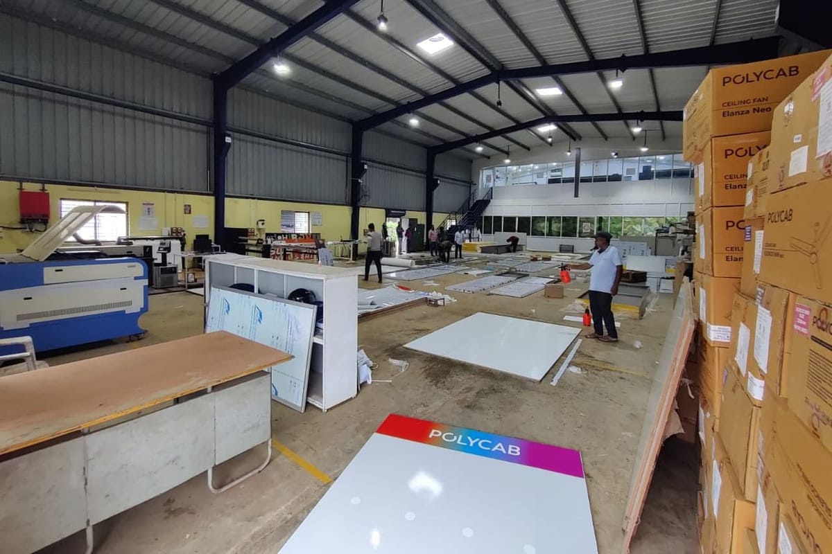 Signage workshop with a Polycab-branded white panel on the floor and stacked Polycab cartons along the right wall